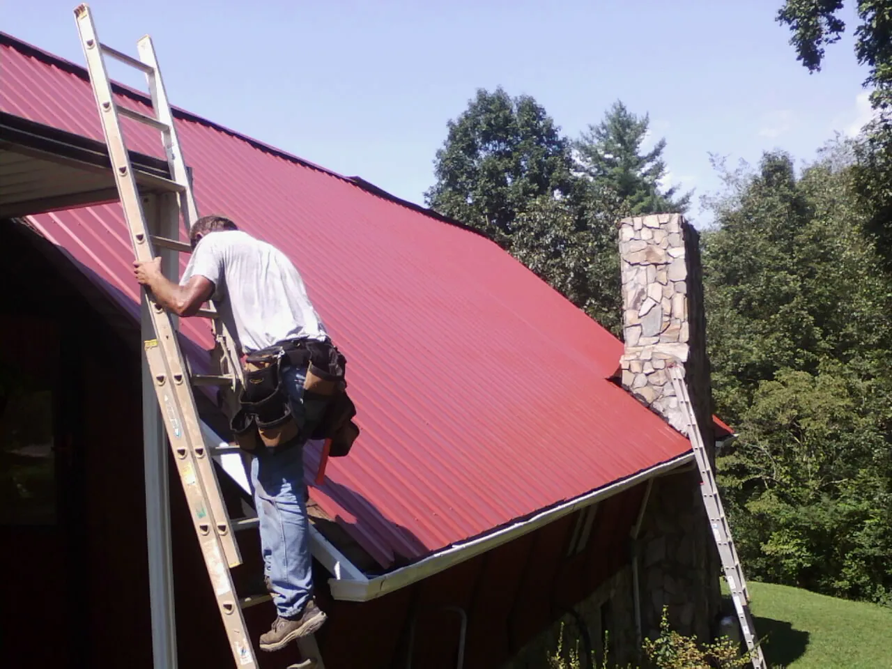 Roofer on a ladder beside a red metal roof and stone chimney — Soffit & Fascia Repair by Roofing Brothers in Saint Donatus
